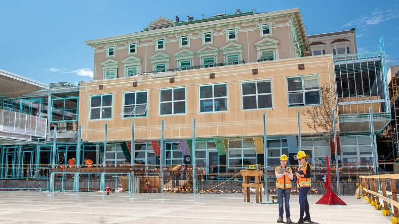 Construction workers look at a tablet in front of a building with a digital overlay of the building design.