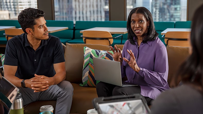 Woman with laptop and man sit on office couch as woman explains