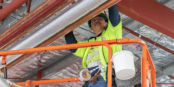 A construction worker on a hydraulic platform lift, working on a set of building pipes