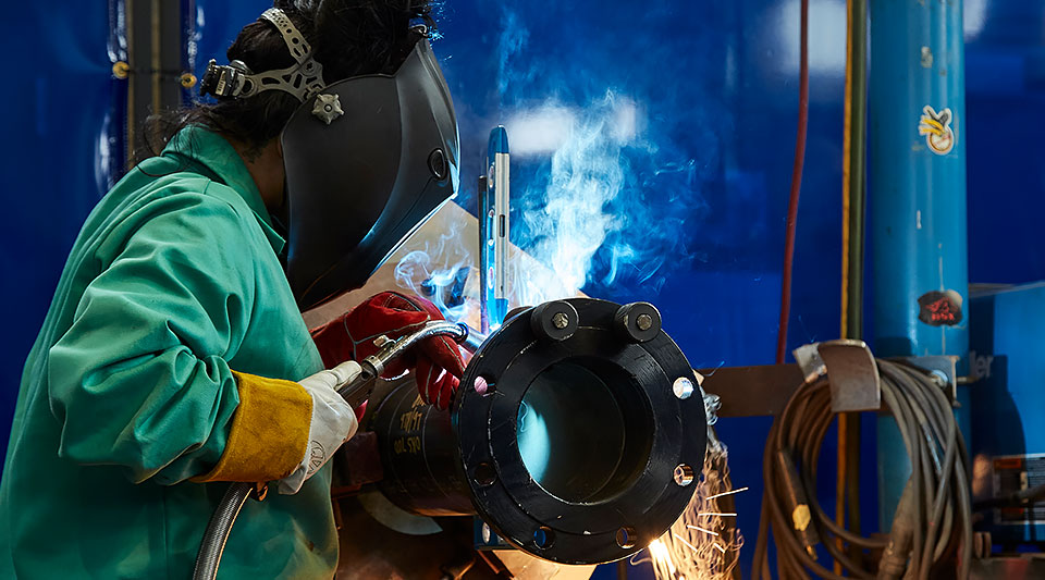 Woman wearing protective welders mask and equipment fabricating a piece of metal piping with sparks flying 