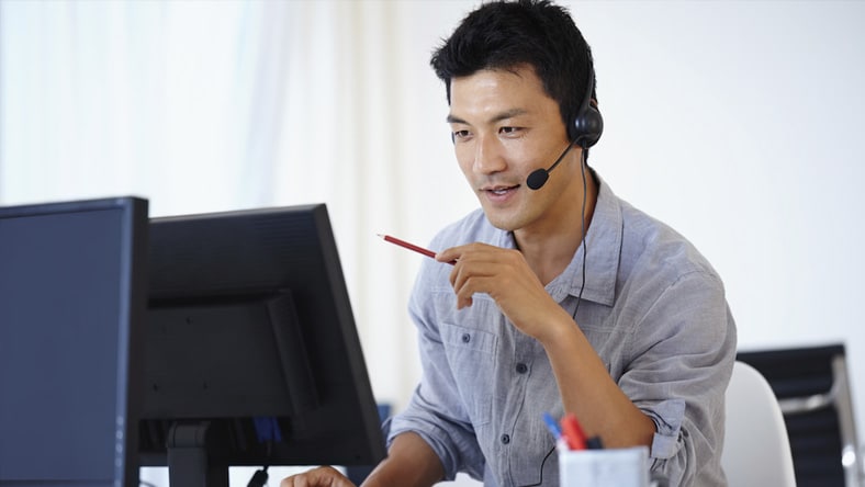 A man in an office looks at his desktop computer screen while talking on a headset.  