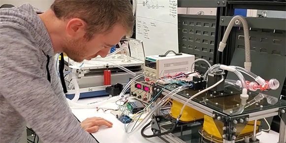 Man in a gray hoodie looking at a ventilator prototype