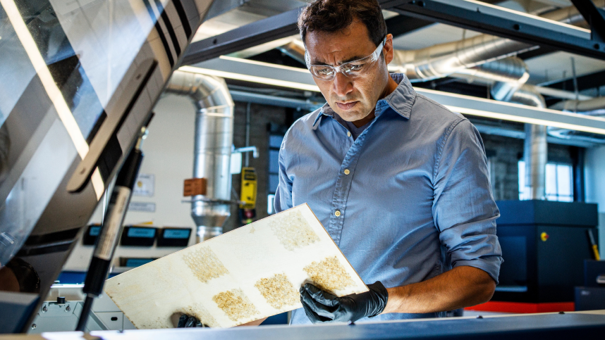 Man in blue shirt and safety glasses surrounded by ductwork, holds machined board 
